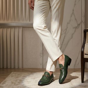 May include: A pair of dark green leather loafers featuring a gold-toned metal horsebit detail across the vamp and a small dark blue accent on the rounded toe. The shoes are worn with slim-fit cream-colored trousers that reach the ankle.