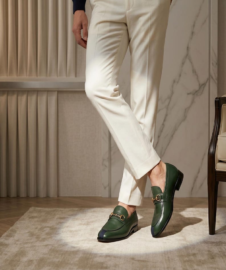 May include: A pair of dark green leather loafers featuring a gold-toned metal horsebit detail across the vamp and a small dark blue accent on the rounded toe. The shoes are worn with slim-fit cream-colored trousers that reach the ankle.