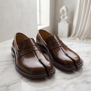 May include: A pair of dark brown leather loafers featuring a distinctive split-toe design. These slip-on shoes have a smooth, polished finish, a traditional penny loafer strap across the vamp, and a low heel with a matching brown sole.