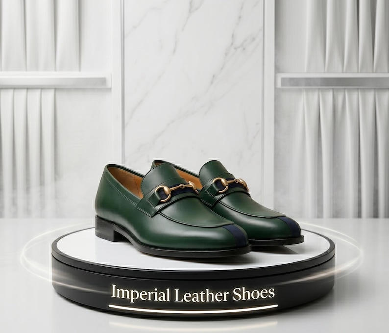 May include: A pair of dark green leather loafers featuring a gold-toned metal horsebit detail across the vamp and a dark blue stripe accent on the toe. The shoes are displayed on a circular stand with the text Imperial Leather Shoes printed on it.
