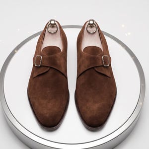May include: A pair of brown suede monk strap shoes on a rotating display. Each shoe has a single strap with a silver buckle. The shoes are presented on a white circular surface.