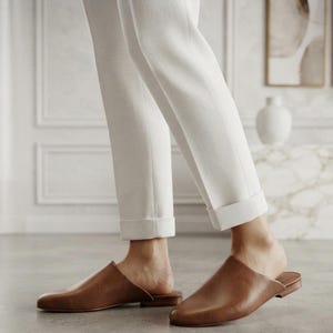 May include: A pair of brown leather mules with a rounded toe and a low heel, worn with white trousers that feature a cuffed hem. The footwear has a smooth finish and a slip-on design, providing a clean and simple look for the lower body.