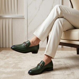 May include: A pair of dark green leather loafers featuring a gold-toned metal horsebit detail across the vamp and a small navy blue accent on the rounded toe. The shoes have a low heel and a smooth finish, worn with cream-colored trousers.