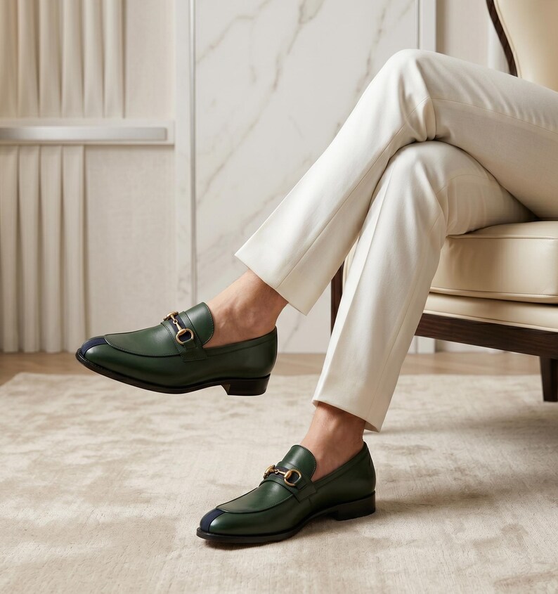 May include: A pair of dark green leather loafers featuring a gold-toned metal horsebit detail across the vamp and a small navy blue accent on the rounded toe. The shoes have a low heel and a smooth finish, worn with cream-colored trousers.