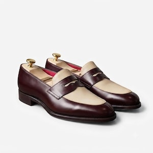 May include: A pair of two-tone penny loafers featuring a deep burgundy leather body and a contrasting cream-colored leather vamp. Each shoe includes a wooden shoe tree with a gold-toned metal knob inserted inside to maintain the shape of the footwear.