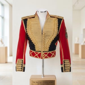 Puede incluir: Una chaqueta de estilo militar roja y negra con adornos trenzados dorados, charreteras y filas de botones dorados. La prenda se exhibe en un soporte con una placa que dice The 'BAD' RED HUSSAR JACKET, MJ COLLECTION, mostrando detalles.