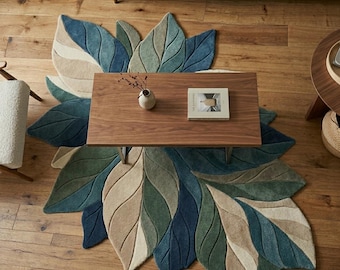 May include: A leaf-shaped area rug in shades of blue, green, and beige, placed on a wooden floor. A rectangular wooden coffee table sits atop the rug, with a small vase and a book on it. The rug's design resembles large leaves.