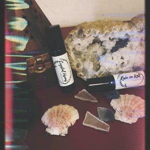 May include: Two black rollerball bottles with white labels featuring the text Crystal Visions and Rain on Art, accompanied by a large geode, two seashells, and three small pieces of sea glass, all arranged together in a collection.