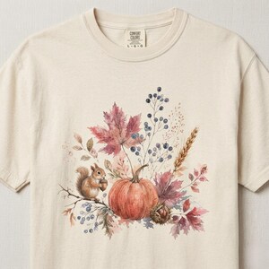 May include: A cream-colored short-sleeve t-shirt featuring a watercolor-style graphic of a brown squirrel holding an acorn, a large orange pumpkin, red maple leaves, wheat stalks, and dark blue berries. The inner neck label reads Comfort Colors L G G.