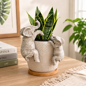May include: A white ceramic planter with a textured geometric pattern, featuring two sculpted grey elephant figures climbing the sides. The planter holds a green snake plant and sits on a circular wooden saucer base on a wooden surface.