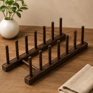 May include: Two dark brown wooden plate racks. Each rack has a rectangular base with vertical pegs for holding plates. The racks are made of stained wood and are designed to hold multiple plates. A small white vase with greenery is in the background.
