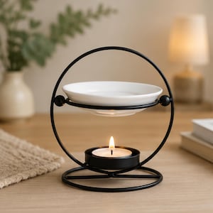 Puede incluir: Un calentador de aceite de aromaterapia de metal negro con un plato de cerámica blanco y una vela de té encendida. El calentador tiene un diseño circular con una base y un anillo superior, y está sobre una superficie de madera.