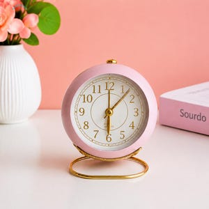 May include: A pink analog alarm clock with a gold-colored base. The clock face is white with black numerals and gold hands. The clock is round and has a gold-colored top. The clock is sitting on a white surface.