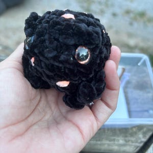 May include: A small, round, black crocheted plush toy with a textured, fuzzy surface. It features two large, glossy, dark brown eyes with a subtle shimmer and small, light pink accents scattered across its body, including a tiny pink tongue.