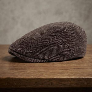 May include: A brown flat cap featuring a textured herringbone pattern. The cap has a rounded crown and a short, stiff brim at the front. The fabric displays a woven appearance with subtle variations in shade, creating a consistent look across the surface.