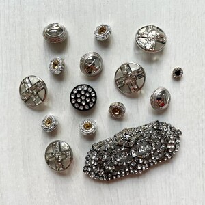 Può includere: Una collezione di bottoni decorativi assortiti e una grande spilla ovale. Gli articoli presentano metallo color argento, cristalli trasparenti e piccole pietre color ambra. I bottoni variano per dimensioni e forma, inclusi design rotondi e a croce.