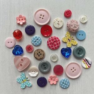 Può includere: Una collezione di bottoni assortiti di varie forme, dimensioni e colori, inclusi design rotondi, a forma di cuore e di farfalla. I bottoni presentano materiali come plastica, legno e tessuto testurizzato in tonalità di rosso, blu, rosa e bianco.