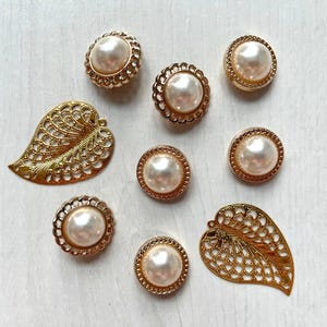 May include: A collection of five round buttons featuring a central white faux pearl surrounded by a gold-toned scalloped metal rim, accompanied by two gold-toned metal leaf-shaped charms with an intricate openwork filigree pattern.
