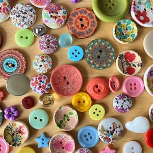 Può includere: Una collezione di bottoni da cucito assortiti in varie dimensioni, forme e colori. I bottoni presentano motivi diversi, tra cui disegni floreali, geometrici e a tinta unita, realizzati in materiali come plastica, legno e metallo, sparsi su una superficie.