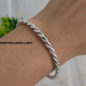 Può includere: Un bracciale color argento caratterizzato da un design a corda intrecciata. Il metallo presenta una finitura lucida e riflettente con un motivo a spirale continuo lungo tutto il cinturino. Il testo KARTHIKJEWELLERY è visibile in nero a sinistra.