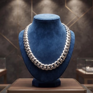 Peut inclure: Un collier à chaîne gourmette épais en métal argenté poli, composé de maillons métalliques arrondis et entrelacés. La chaîne robuste présente une finition métallique brillante et réfléchissante sur un buste de présentation en velours bleu.