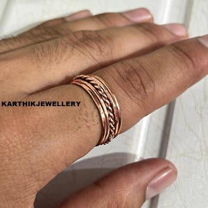 May include: A copper ring featuring a central twisted rope-style band flanked by two smooth, flat bands on either side. The ring has a warm, metallic reddish-brown finish. The text KARTHIKJEWELLERY is visible on the left side of the image.