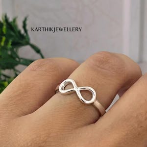 May include: A silver-toned ring featuring an infinity symbol design on the band. The text KARTHIKJEWELLERY is visible in the upper portion of the frame. The ring has a polished metallic finish and a continuous loop shape representing infinity.