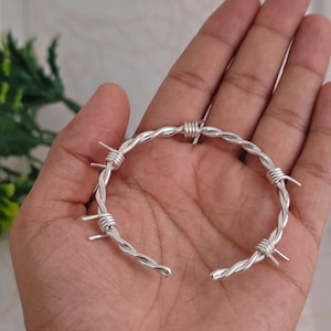 Puede incluir: Una pulsera tipo brazalete de tono plateado diseñada para parecerse a un trozo de alambre de espino. La banda metálica presenta una construcción de alambre trenzado con varias púas sobresalientes espaciadas uniformemente alrededor del marco.
