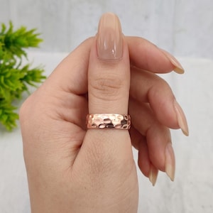 Puede incluir: Un anillo de banda en oro rosa que presenta una textura martillada en toda su superficie exterior. El metal tiene un acabado pulido y reflectante que capta la luz, creando una serie de pequeñas hendiduras cóncavas alrededor del anillo.