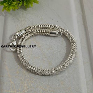 Op de afbeelding: Een zilverkleurige kettingarmband met een geweven, gevlochten schakelpatroon. De armband is voorzien van een karabijnsluiting en een ronde ring aan de uiteinden. De tekst KARTHIKJEWELLERY staat in het zwart aan de linkerkant van de armband.