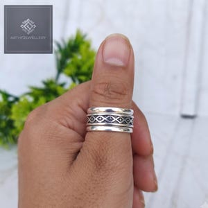 Pode incluir: Um anel de prata com uma banda central apresentando um padrão repetitivo de diamantes e estrelas, ladeado por duas bandas de prata lisas. O metal tem um acabamento polido e os detalhes gravados são realçados com oxidação escura.