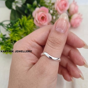 Pode incluir: Um anel de prata polida com um design de banda lisa e ondulada. O anel é usado num polegar, e o texto KARTHIK JEWELLERY está impresso a preto no lado esquerdo da imagem, posicionado perto do fundo floral cor-de-rosa desfocado.