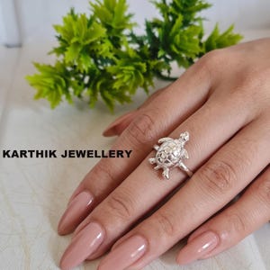 Puede incluir: Un anillo de tono plateado con un diseño detallado de tortuga en la banda. La tortuga tiene un caparazón texturizado y cuatro aletas visibles. El texto KARTHIK JEWELLERY está impreso en negro a la izquierda de la imagen, junto al anillo.