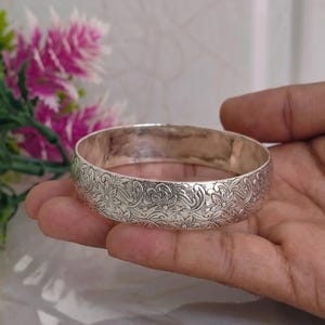 Puede incluir: Una pulsera rígida de color plateado que presenta un patrón continuo de flores y volutas en relieve en toda la superficie exterior. La banda de metal tiene un acabado pulido y un perfil redondeado con detalles botánicos intrincados.