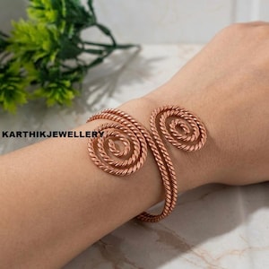 May include: A copper-colored metal cuff bracelet featuring two prominent spiral designs made of twisted wire. The bracelet has an open-ended structure that wraps around the wrist. The text KARTHIKJEWELLERY is visible on the left side of the image.
