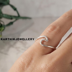 Puede incluir: Un anillo de plata con un diseño continuo en forma de onda en la banda. El texto KARTHIKJEWELLERY está impreso en negro en el lado izquierdo de la imagen. El anillo se lleva en un dedo, mostrando el acabado metálico liso y pulido de la pieza.