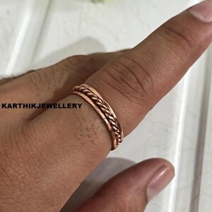 Puede incluir: Un anillo de cobre que presenta un diseño central de cuerda trenzada flanqueado por dos bandas lisas. El anillo tiene un acabado metálico marrón rojizo y se lleva en un dedo. El texto KARTHIKJEWELLERY es visible en el lado izquierdo.