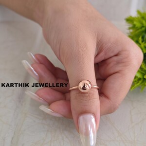 May include: A rose gold ring featuring a single polished spherical bead at the centre. The band is thin and circular, showcasing a smooth metallic finish. The text KARTHIK JEWELLERY is visible to the left of the ring on the light background.