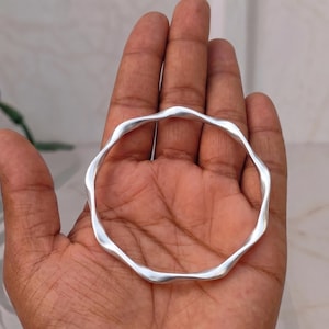 May include: A silver-toned bangle bracelet featuring a continuous, smooth, wavy or undulating edge design. The circular accessory has a polished, reflective metallic surface with a consistent thickness throughout its entire circumference.