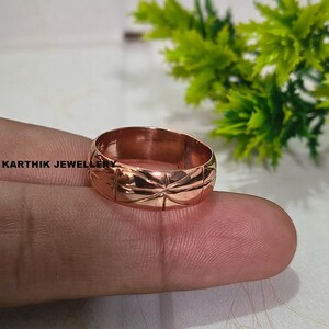 Può includere: Un anello in rame lucido caratterizzato da un motivo geometrico a stella ripetuto inciso attorno alla fascia esterna. Il metallo ha una calda lucentezza metallica bruno-rossastra. Il testo KARTHIK JEWELLERY è visibile in nero sulla sinistra.