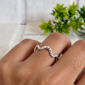 Puede incluir: Un anillo de plata con una textura de cuerda trenzada y un diseño ondulado. El metal tiene un acabado pulido y reflectante. El texto (KARTHIKJEWELLERY) es visible en el fondo. El anillo se lleva puesto en un dedo, mostrando su forma.
