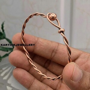 Puede incluir: Una pulsera de metal color cobre con un diseño de alambre trenzado. La pulsera tiene forma circular con una cuenta esférica pulida en un extremo, que sirve como terminal decorativo. El metal presenta un acabado metálico cálido.