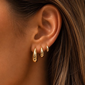 Peut inclure: Trois créoles dorées de tailles différentes disposées en rangée sur le lobe de l'oreille. Chaque boucle d'oreille présente un design lisse, poli et en forme de goutte d'eau avec une finition métallique réfléchissante, créant un aspect gradué.