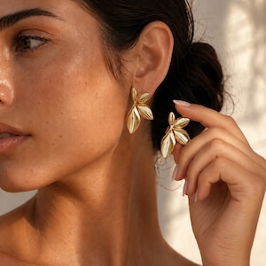 May include: A pair of gold-toned earrings featuring a cluster of three smooth, polished leaf-shaped elements. The earrings have a metallic finish and are designed with a layered, botanical-inspired arrangement that catches the light.