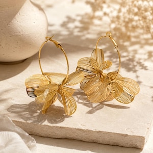 Pode incluir: Um par de brincos de argola em tom dourado com pétalas de arame em camadas que criam uma forma floral. Os brincos são construídos com um fio de metal dourado fino disposto num padrão delicado e aberto que imita a aparência de flores desabrochadas.