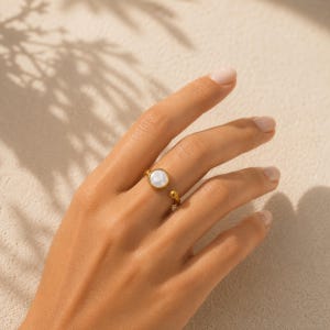 May include: A gold-toned open ring featuring a circular white mother-of-pearl inlay on one end and a small gold-toned sphere on the other. The band is thin and polished, showcasing a simple design that wraps around the finger with an open gap.