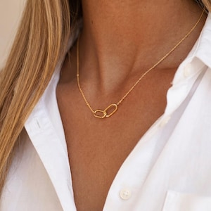 Peut inclure: Un collier doré composé d'une chaîne fine et d'un pendentif central formé de deux maillons ovales entrelacés. Le collier repose sur une chemise blanche à col, mettant en valeur une finition métallique polie et un design simple et épuré.