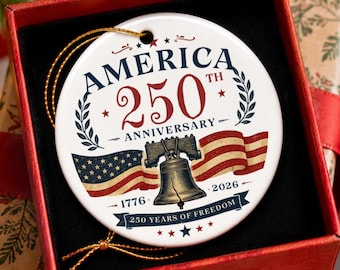 May include: A round white ceramic ornament featuring a graphic of the Liberty Bell, the American flag, and the text AMERICA 250TH ANNIVERSARY 1776 2026 250 YEARS OF FREEDOM. The ornament has a gold hanging string and is presented in a red gift box.