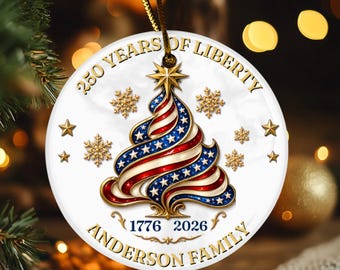 May include: A round white ornament featuring a stylized Christmas tree with red, white, and blue stripes and stars. The text reads 250 YEARS OF LIBERTY, 1776 2026, and ANDERSON FAMILY. Gold stars and snowflakes surround the tree, suspended by a gold cord.