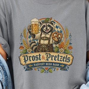 Puede incluir: Una camiseta gris con un gráfico de un mapache vestido con ropa tradicional bávara, sosteniendo una jarra de cerveza y un pretzel. El texto dice Prost & Pretzels Harvest Beer Bash. El diseño incluye tallos de trigo y patrones de rombos azules.